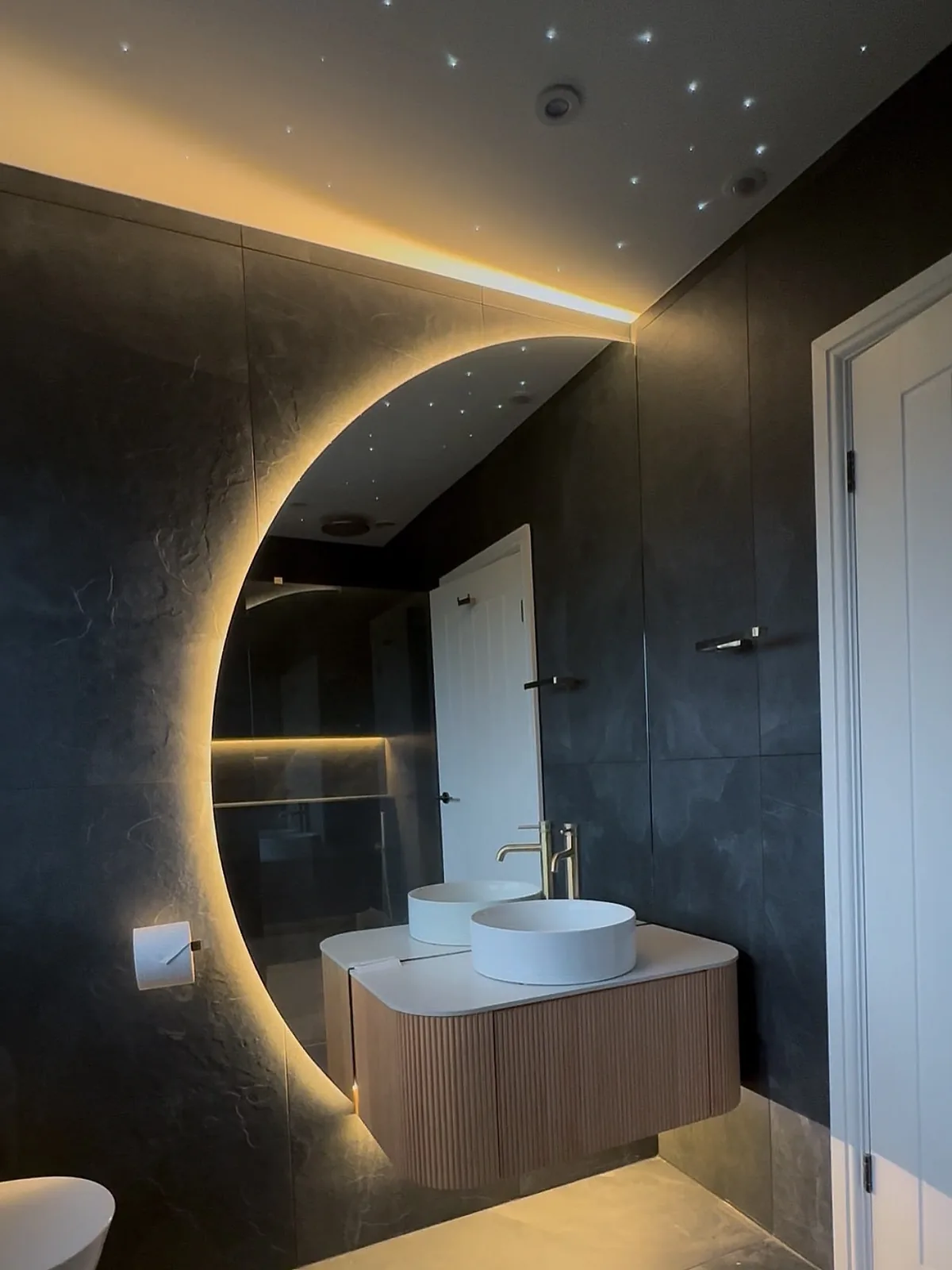 Dark tiled bathroom renovation with slatted timber vanity, vessel basin and crescent backlit mirror