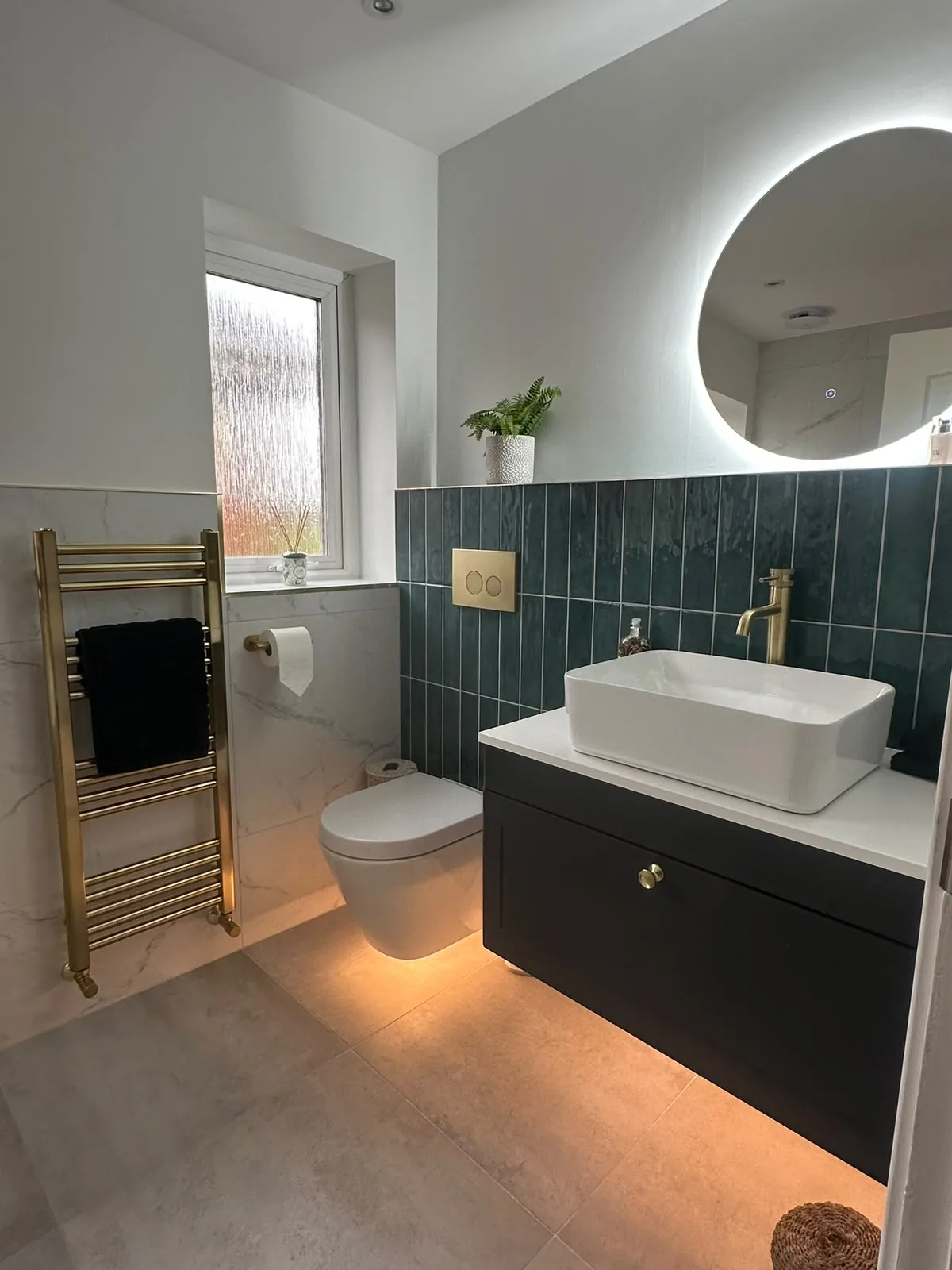 Small bathroom renovation with green vertical tiles, wall-hung toilet, black vanity and backlit round mirror